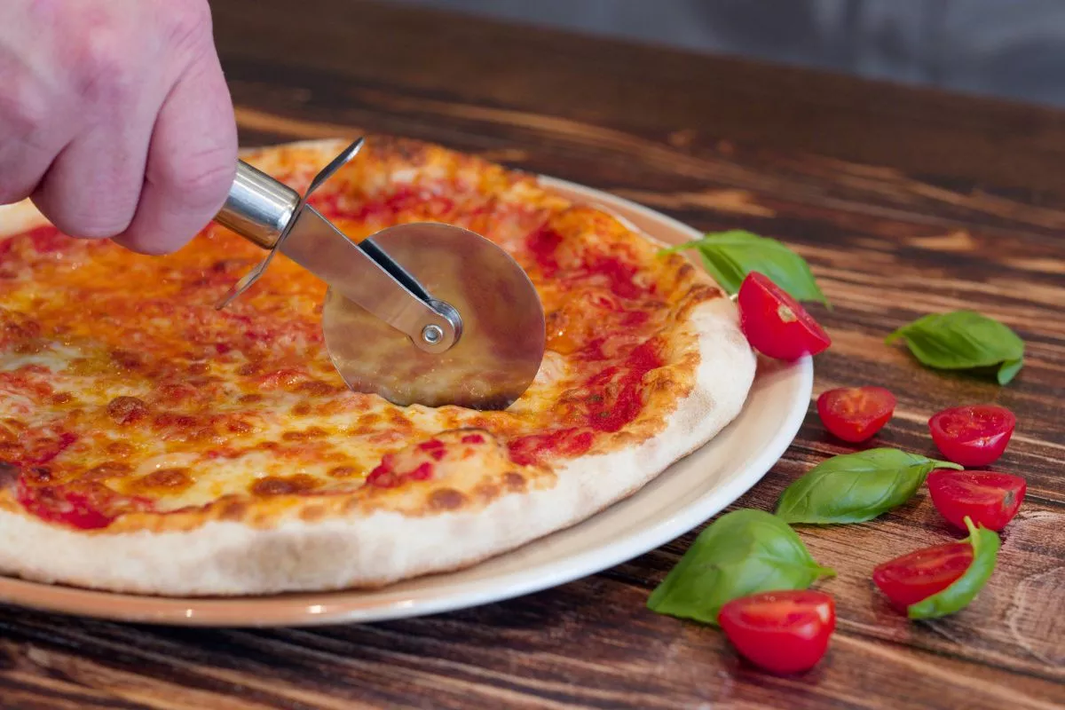 Leckere Pizza Margharita, die gerade mit einem Pizzaschneider geschnitten wird.